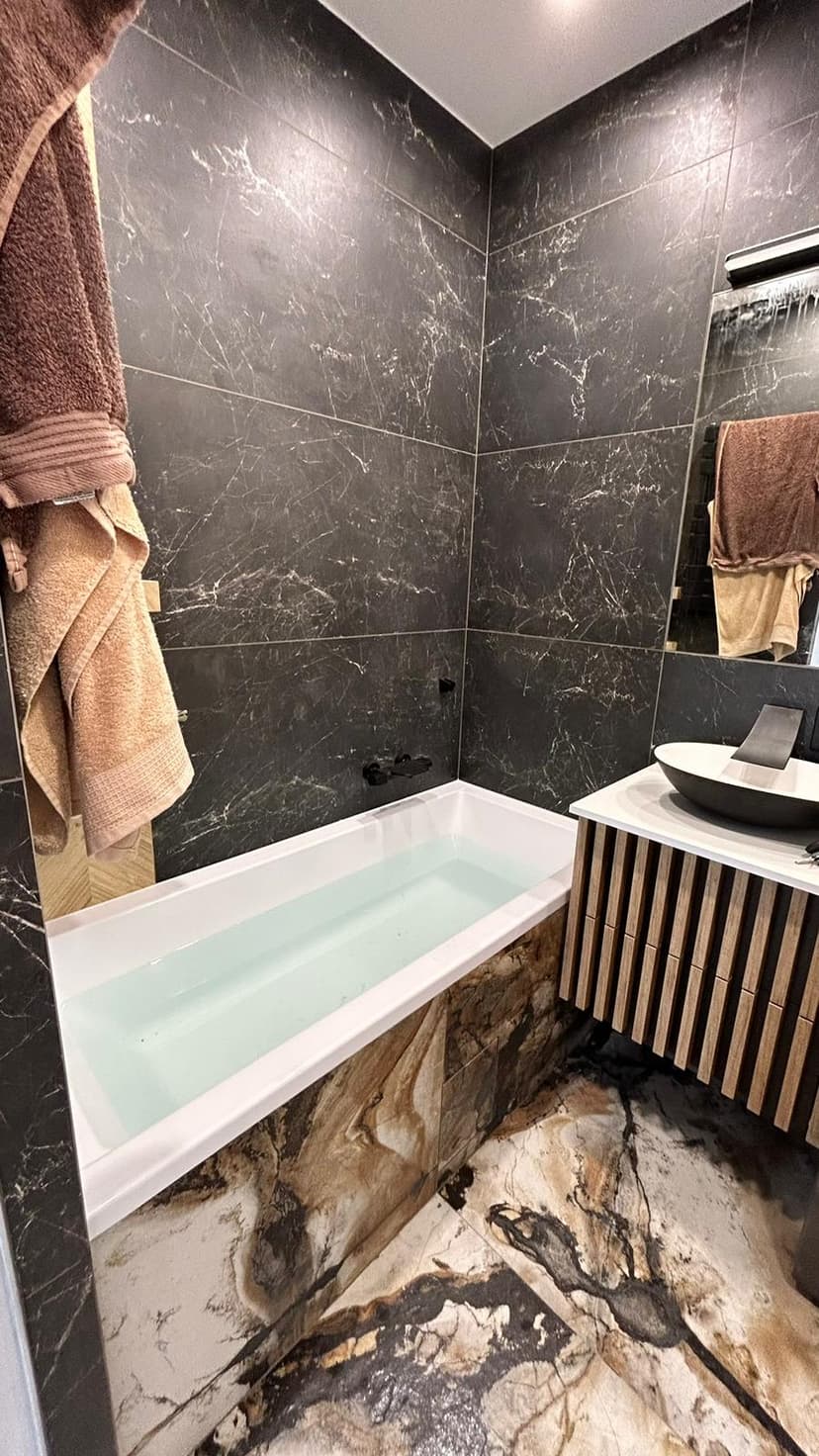 Łazienka w płytki o granitowej kolorystyce i fakturze marmuru, szafka łazienkowa o strukturze drewna i ocieplające wnętrze brązowo-kremowe tekstylia.