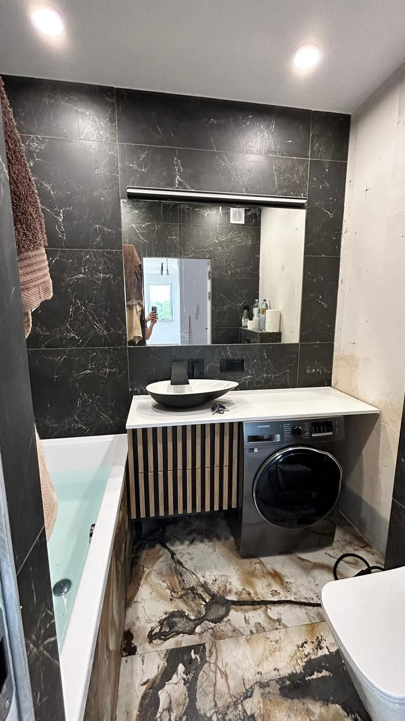 Łazienka w płytki o granitowej kolorystyce, zabudowa umywalki i ukryta pod blatem umywali pralka, nad umywalką duże lustro.