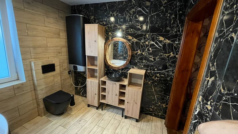 Łazienka z antracytową kabiną prysznicową, zabudowa umywalki w kolorze jasnego drewna, czarna terma oraz czarna umywalka i czarny sedes.