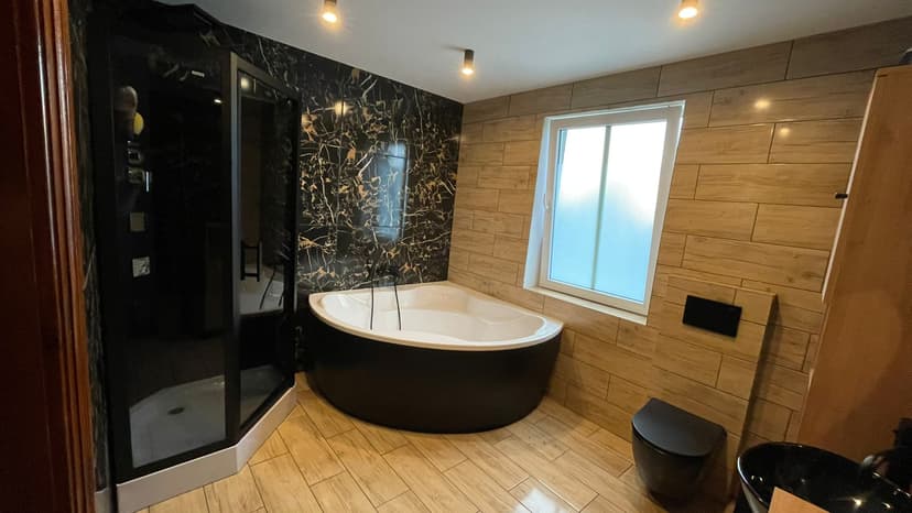 Łazienka z antracytową kabiną prysznicową, jasna ściana z oknem i duża narożna wanna z czarnym wykończeniem i białym środkiem.