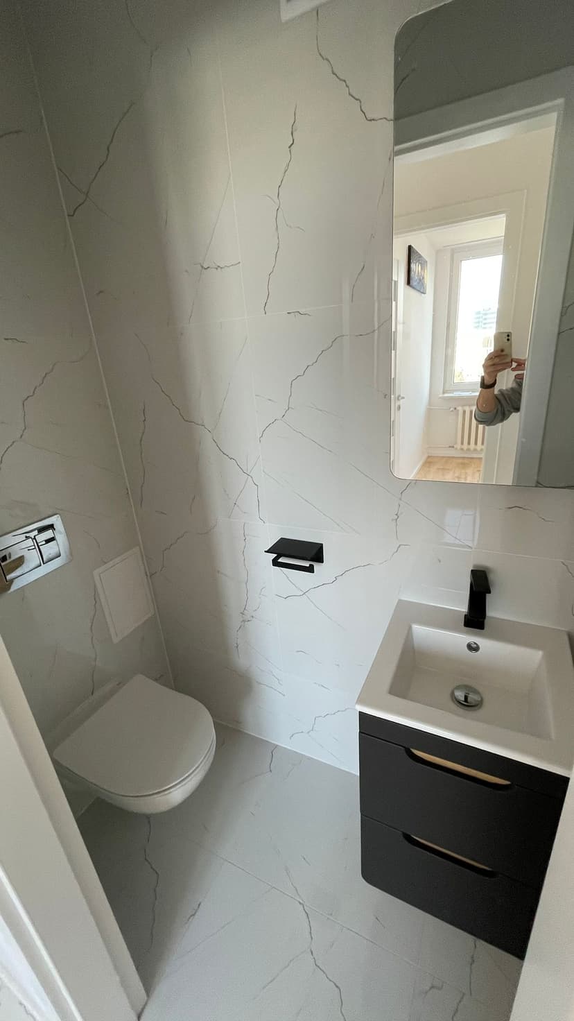 Toaleta w białym marmurze, czarna szafka pod umywalką, czarna armatura i uchwyt na papier toaletowy, duże lustro.