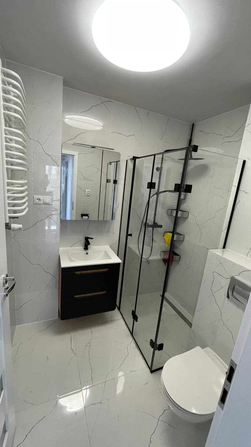 Łazienka w białym marmurze, szklany prysznic typu `walk-in`, czarna szafka pod umywalką, czarna armatura, duże lustro nad szafką.