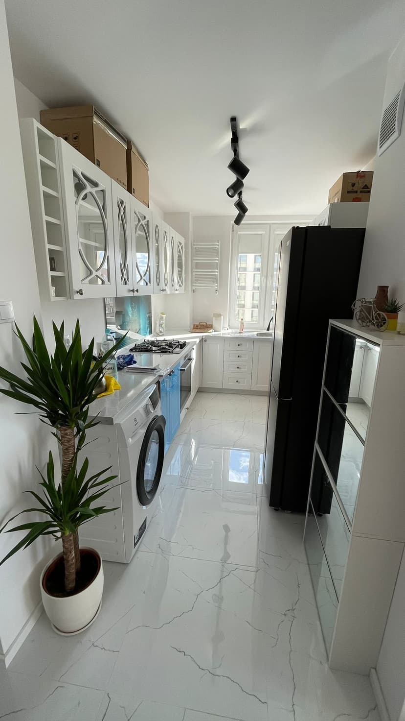 Biała, jasna kuchnia, jasne płytki z fakturą białego marmuru, oszklona biała stolarka kuchenna, czarna lodówka oraz antracytowe oświetlenie sufitowe.