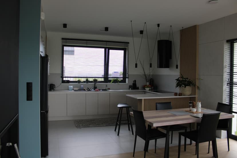 Jasna, nowoczesna kuchnia z półwyspem, modernistycznym okapem, czarnymi elementami oświetlenia, połączona z jadalnią w stylu loftowym z czarnymi krzesłami.