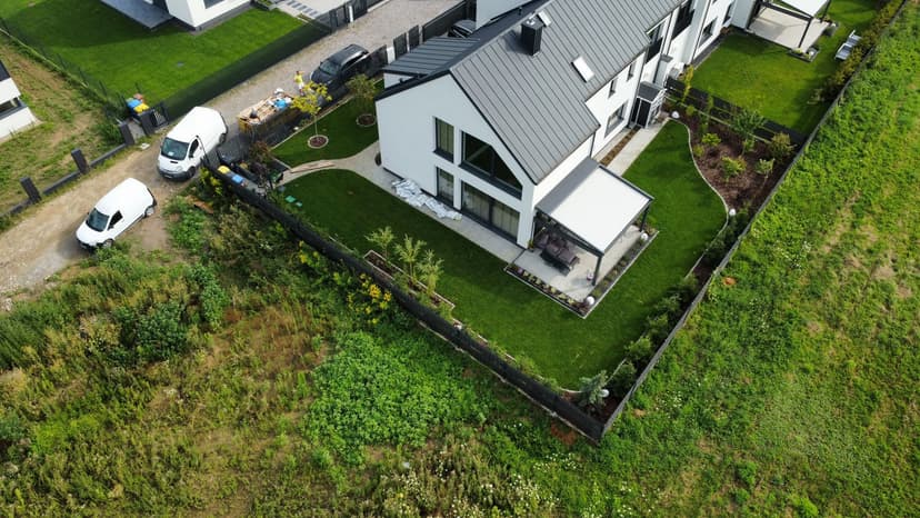 Dom w zabudowie typu bliźniak, biała elewacja, kolor dachu ciemny grafit, zielony trawnik i drzewa przed posesją.