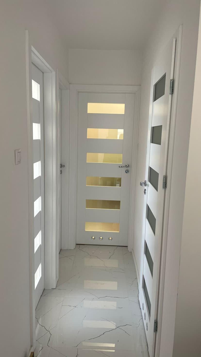 Jasny, biały hol z białymi płytkami podłogowymi o fakturze marmuru, na wprost drzwi wejściowe do łazienki, drzwi ze srebrnymi okuciami oraz białe ościeżnice.