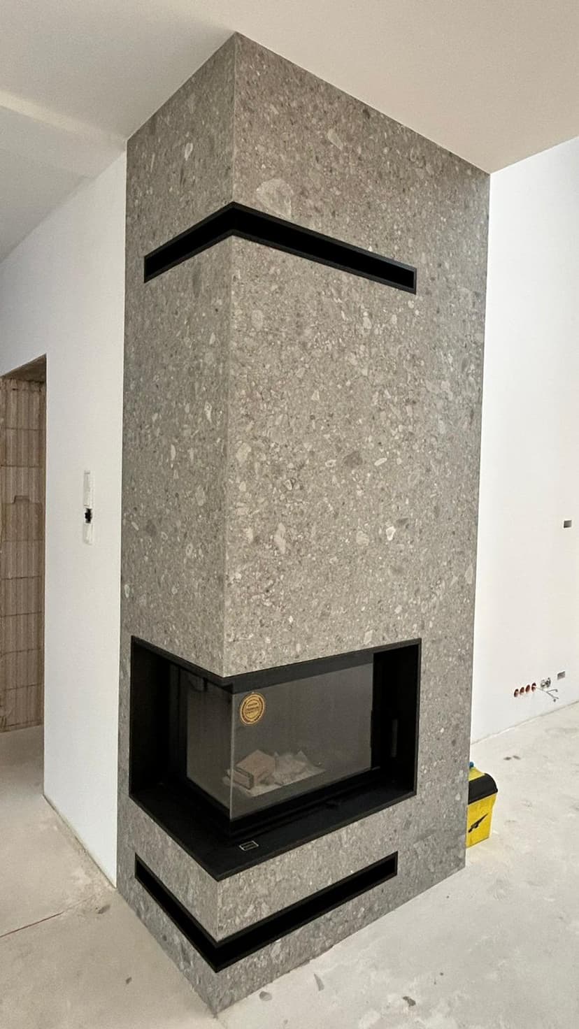 Granitowy kominek narożny z czarnymi okuciami i szybą, poziome wyciągi z czarnymi detalami.