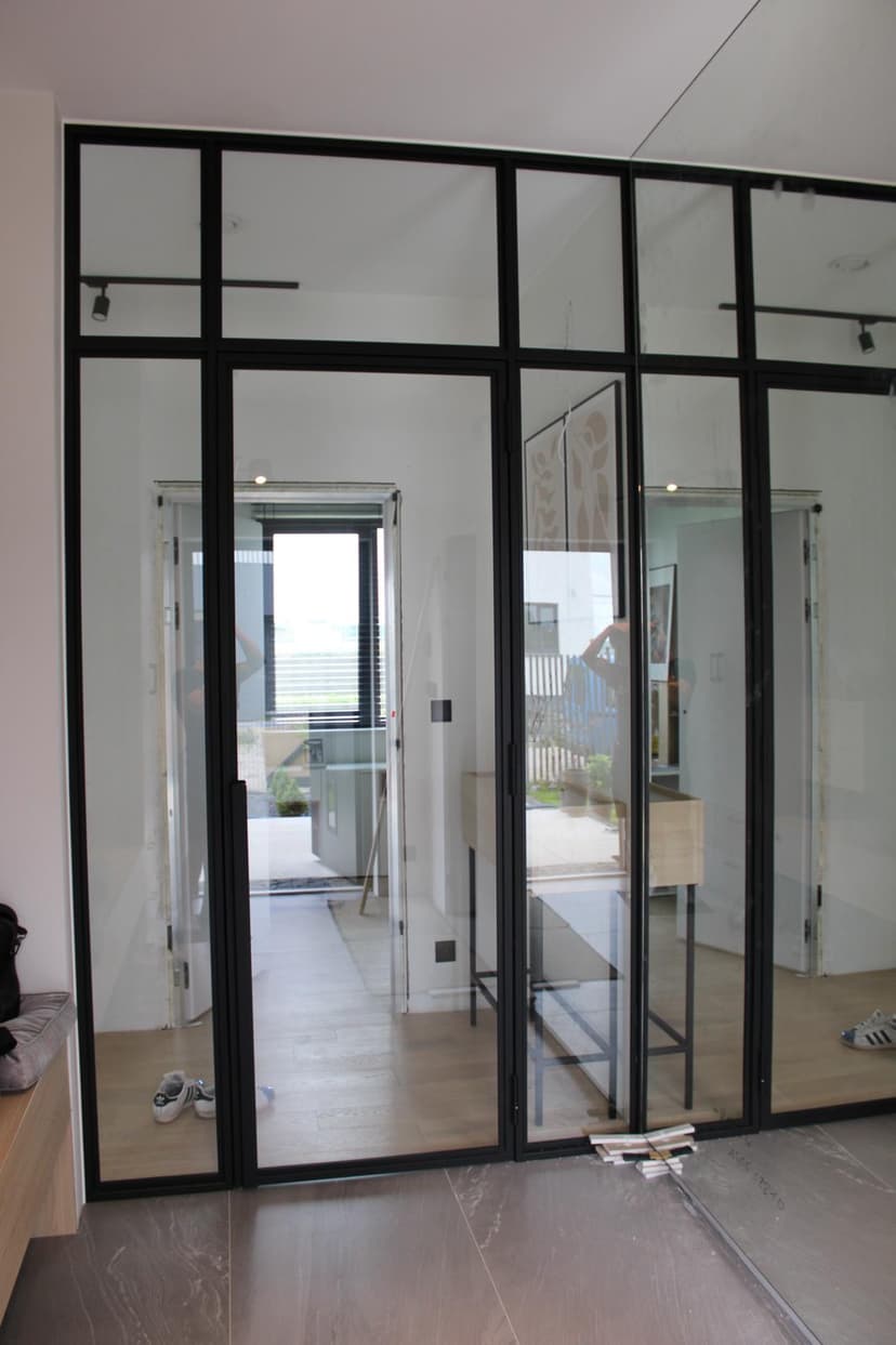 Korytarz prowadzący do wiatrołapu drzwi wejściowych, oszklona zabudowa z czarnymi metalowymi elementami oraz lustro na całą wysokość ściany po prawej stronie.