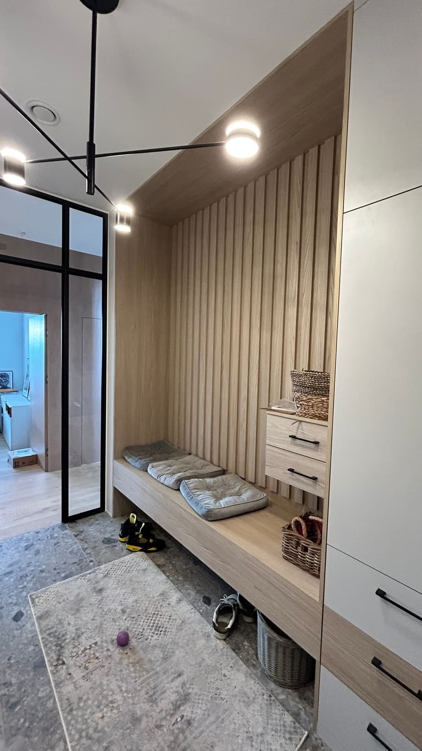 Wyjście z korytarza wiatrołapu do przestrzeni mieszkalnej, miejsce na spoczynek i miejsce na buty, stolarka drewniana w kolorze jasnym, poduszki szare i modernistyczny żyrandol w kolorze czarnym.