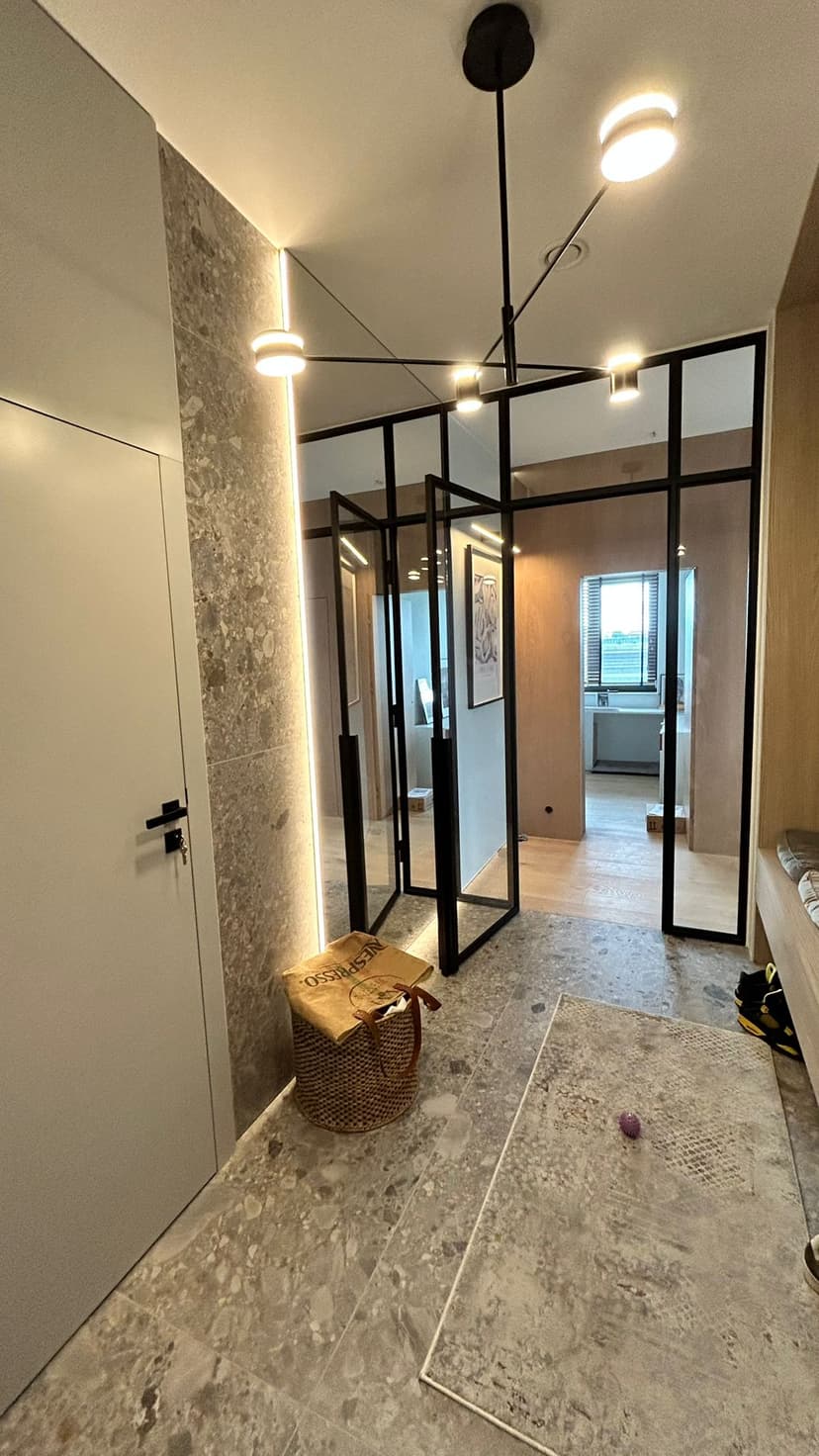 Wyjście z korytarza, na wprost przestrzeń mieszkalna z pokojem typu gabinet, oświetlenie sufitowe w kolorze czarnym, na podłodze oraz ścianie z dużym lustrem kamienne płytki w kolorze jasny granit.