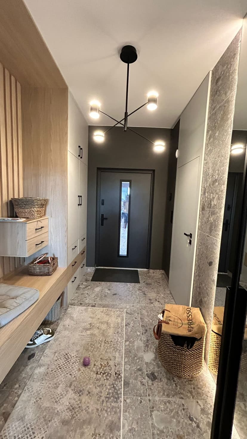 Widok na wyjście z korytarza na drzwi wejściowe, po lewej stronie zabudowa szafek i przechowywanie garderoby, po prawej przejście do garażu i duże lustro, ściana oraz drzwi wejściowe w oddali w kolorze czarnym.