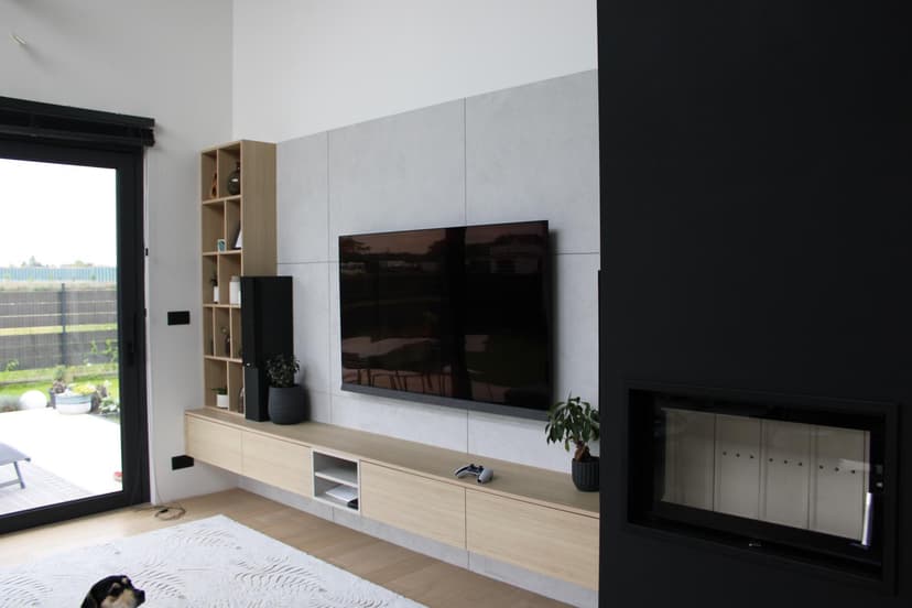 Nowoczesny pokój wypoczynkowy z zabudową, wielkoformatowe jasnoszare płytki na ścianie, podwieszana szafka, podwieszany telewizor LCD z ukrytym w ścianie okablowaniem, jasna stolarka drewniana oraz ciemny narożnik z kominkiem dekoracyjnym, jasny pokój z widokiem na ogród.
