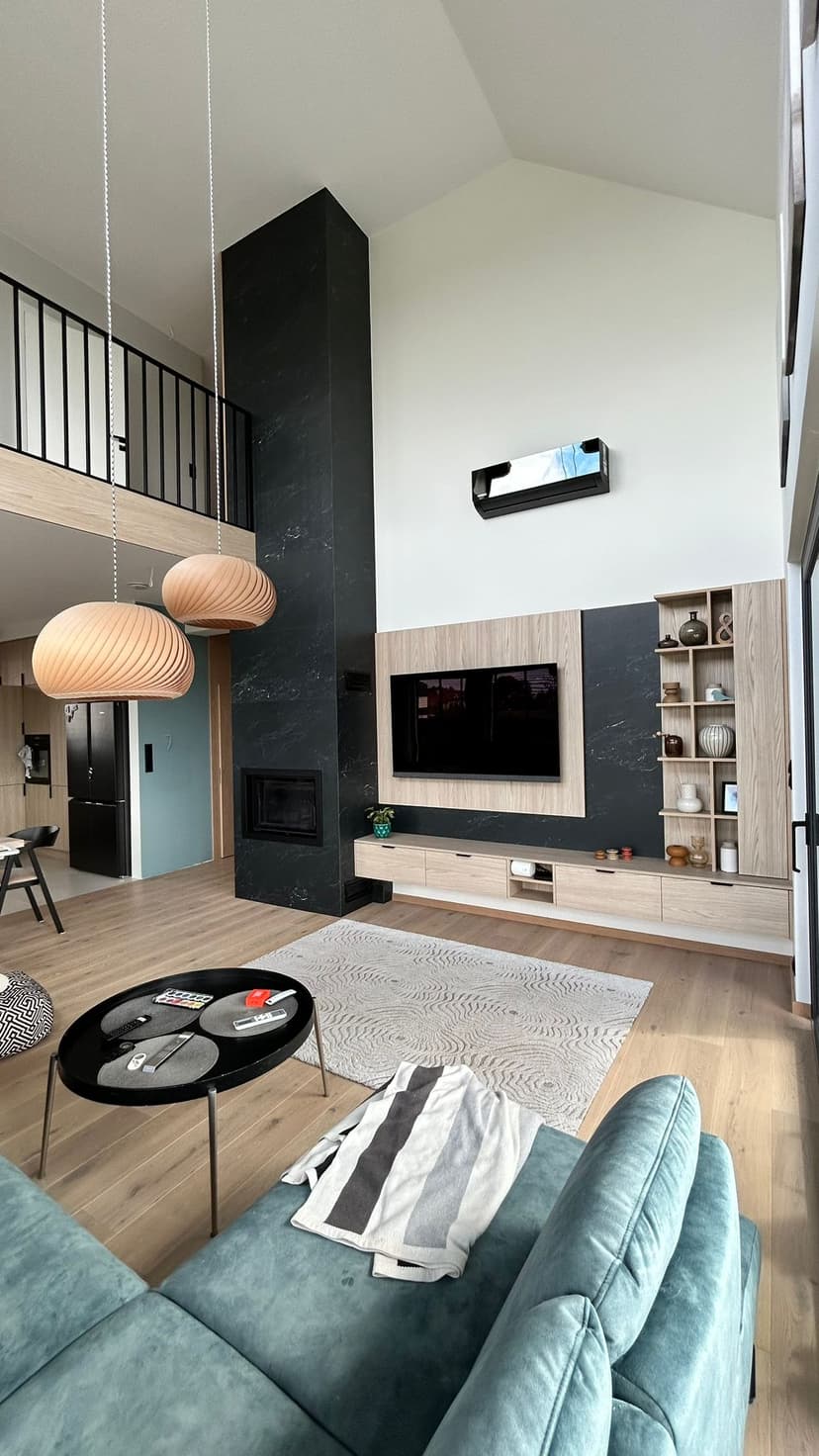 Nowoczesny pokój wypoczynkowy z otwartą przestrzenią, widoczna antresola, czarny kominek, czarny klimatyzator, jasne drewniane parkiety oraz białe ściany.