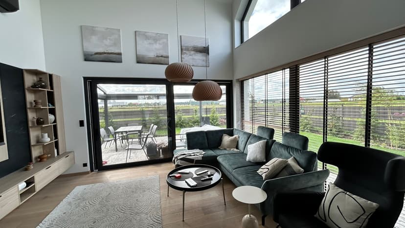 Nowoczesny pokój wypoczynkowy z widokiem na taras i ogród, nowoczesne drewniane rolety oraz antracytowe meble - okrągły stolik kawowy, fotel i zestaw wypoczynkowy, na ścianie tryptyk trzech obrazów z pejzażem.