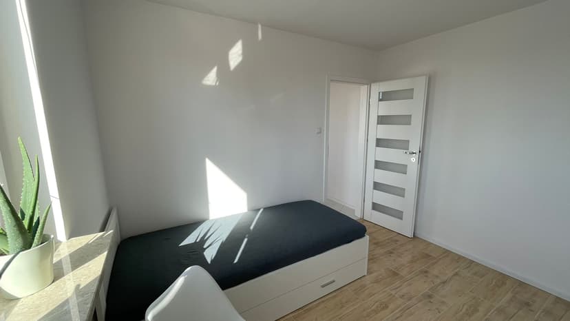 Jasna sypialnia, łóżko jednoosobowe z szufladą do przechowywania pod spodem, jasny pokój z roślinami na parapecie, w tle drzwi wejściowe na korytarz mieszkania.