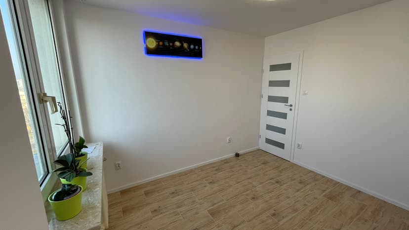 Jasna sypialnia z dużym oknem, jasny parkiet oraz układ planetarny w formie podświetlanego LED obrazu, białe ściany i rośliny na parapecie.